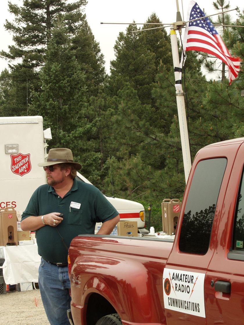 A man in a hat and sunglasses holds a handheld radio beside a red pickup marked Amateur Radio Communications. Behind him stand an antenna mast with a US flag, pine trees and a Salvation Army canteen trailer.
