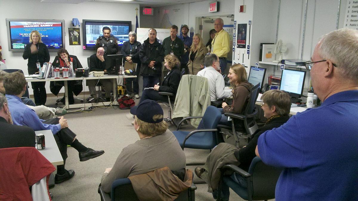 A crowded emergency operations center: about twenty people sit and stand among desks with computer monitors, facing a briefing at the front of the room, with two wall-mounted TV screens showing news coverage.