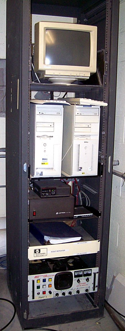 A tall black equipment rack with a CRT monitor on top, two desktop computers below it, then a small radio, a power supply, a binder on a shelf, a panel labeled K2GXT repeater and the repeater unit at the bottom.