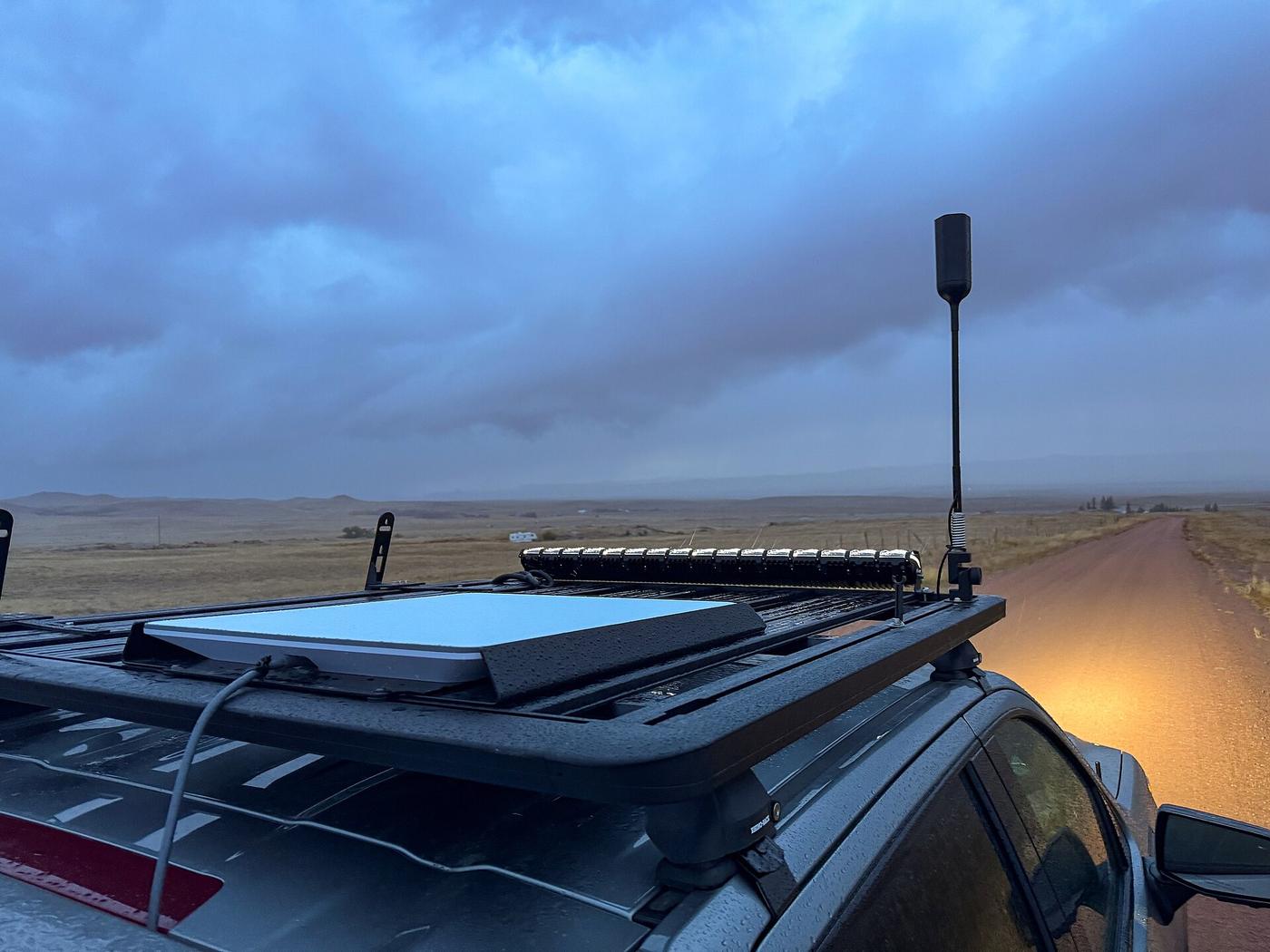 A white rectangular Starlink flat-panel dish mounted on the roof rack of a dark vehicle next to a light bar and a small antenna, under heavy gray storm clouds on an open plain beside a gravel road.